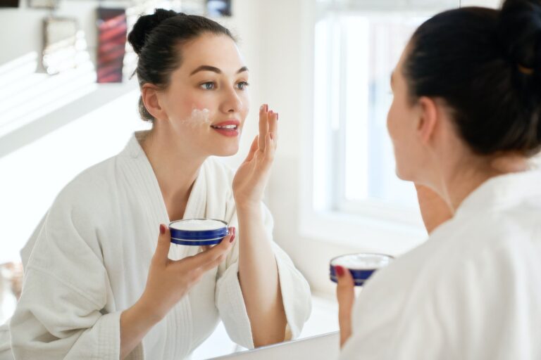 Hermosa mujer joven con crema para la cara mirando en el espejo en el baño en casa. Tratamiento de belleza facial. Cosmetología, belleza y spa.