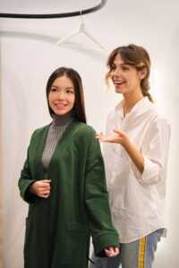 Joven asesora de moda ayudando alegremente a probarse la chaqueta de punto a una hermosa chica asiática en la tienda